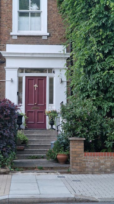 The image depicts the entrance to a residential property with a traditional brick exterior and a prominent white decorative doorframe. The door is painted a deep red or burgundy and features a brass m