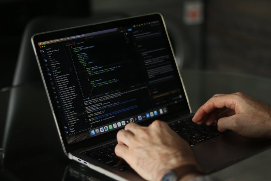 A close-up view of a person's hand typing on a silver laptop keyboard placed on a wooden desk. The laptop screen displays lines of HTML or CSS code, suggesting work related to web development or conte